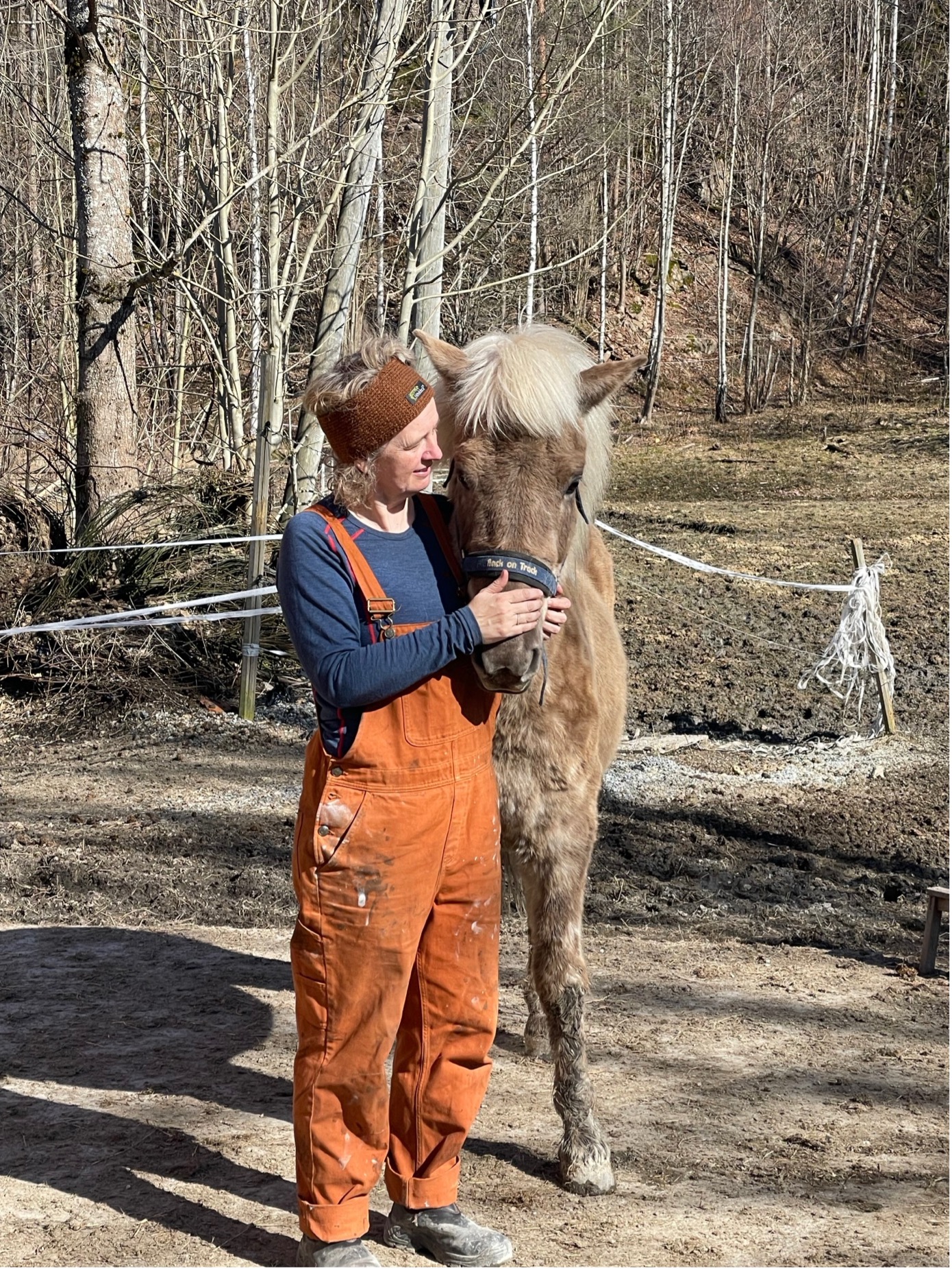 Kvinne i oransje kjeledress som står tett inntil en lys hest